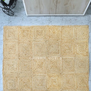 Puede incluir: Una alfombra de fibra natural con un patr&oacute;n cuadrado. La alfombra est&aacute; hecha de yute tejido y tiene un color beige claro. La alfombra est&aacute; compuesta por una serie de cuadrados, cada uno con una textura ligeramente diferente. La alfombra es perfecta para agregar un toque de belleza natural a cualquier habitaci&oacute;n.