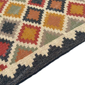 May include: A close-up of a woven rug with a geometric pattern in shades of brown, red, green, and black. The rug has a fringe border.