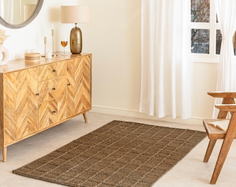 May include: A rectangular brown jute rug with a diamond pattern sits on a light-colored floor. The rug is in a room with a wooden cabinet, a lamp, a mirror, and a wooden chair. The room has white curtains and a window.