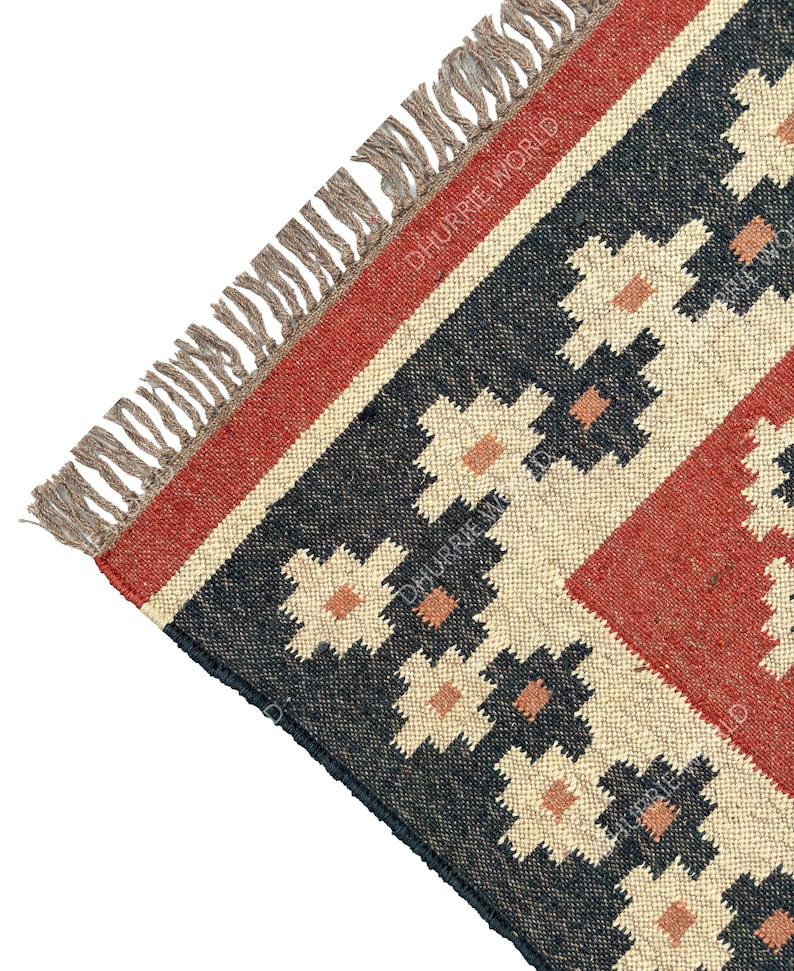 May include: Close-up of a corner of a black, red, and beige patterned rug with fringe. The rug has a geometric design with floral motifs. The text "DHURRIE WORLD" is visible in the image.