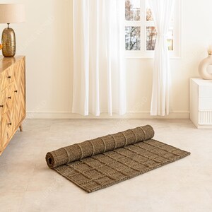 Puede incluir: Una alfombra rectangular marr&oacute;n enrollada con un patr&oacute;n tejido en forma de cuadr&iacute;cula. La alfombra est&aacute; parcialmente desenrollada sobre un suelo de color claro. La habitaci&oacute;n tiene una paleta de colores neutros con una c&oacute;moda de madera y cortinas blancas en el fondo.