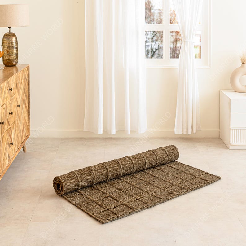 Puede incluir: Una alfombra rectangular marr&oacute;n enrollada con un patr&oacute;n tejido en forma de cuadr&iacute;cula. La alfombra est&aacute; parcialmente desenrollada sobre un suelo de color claro. La habitaci&oacute;n tiene una paleta de colores neutros con una c&oacute;moda de madera y cortinas blancas en el fondo.