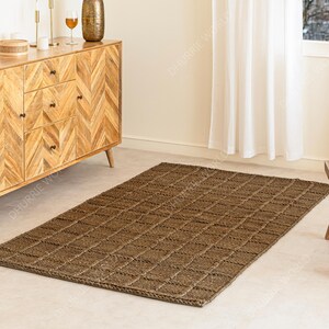 Puede incluir: Una alfombra rectangular de yute marr&oacute;n con un patr&oacute;n tejido en forma de cuadr&iacute;cula. La alfombra est&aacute; colocada sobre un suelo de color claro, con un mueble de madera y una ventana con cortinas blancas en el fondo. Es un art&iacute;culo de decoraci&oacute;n del hogar.