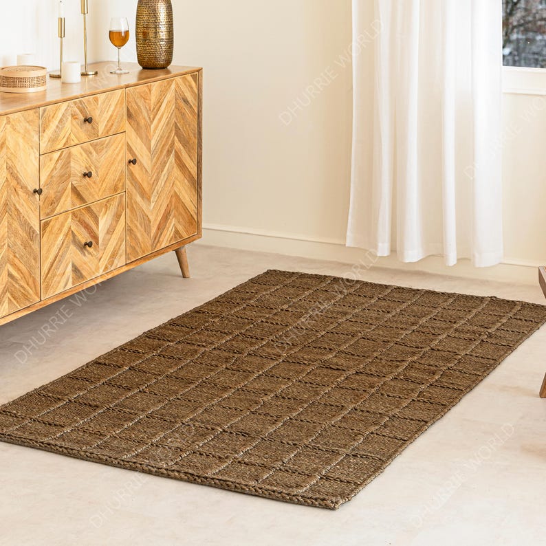 Puede incluir: Una alfombra rectangular de yute marr&oacute;n con un patr&oacute;n tejido en forma de cuadr&iacute;cula. La alfombra est&aacute; colocada sobre un suelo de color claro, con un mueble de madera y una ventana con cortinas blancas en el fondo. Es un art&iacute;culo de decoraci&oacute;n del hogar.