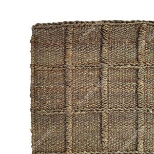 Puede incluir: Primer plano de una alfombra tejida con un patr&oacute;n texturizado en forma de cuadr&iacute;cula. La alfombra est&aacute; hecha de fibras naturales en tonos marrones y beige. Los bordes est&aacute;n cuidadosamente acabados, mostrando una est&eacute;tica r&uacute;stica y artesanal.