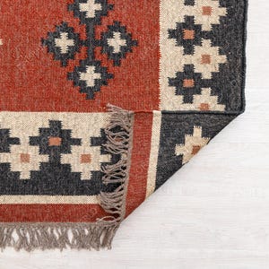 May include: A red, black, and cream colored rug with a geometric pattern. The rug has fringe on all four sides and is folded in half.