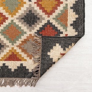 May include: A close-up of the corner of a woven rug with a geometric pattern in shades of brown, red, yellow, and green. The rug has a black border and fringe along the edges.