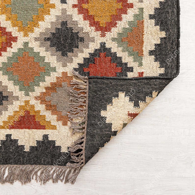 May include: A close-up of the corner of a woven rug with a geometric pattern in shades of brown, red, yellow, and green. The rug has a black border and fringe along the edges.