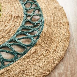 May include: Round jute rug with a teal border. The rug has a natural, woven texture with a teal rope-like design around the edge. The rug is on a light wood floor.