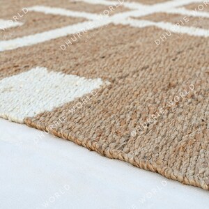 May include: A close-up of a natural fiber rug with a white geometric pattern. The rug is made of jute and has a natural, earthy color.