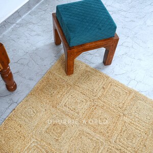Puede incluir: Un coj&iacute;n de terciopelo turquesa se sienta sobre un taburete de madera con una alfombra de yute natural debajo. La alfombra tiene un patr&oacute;n tejido de cuadrados.