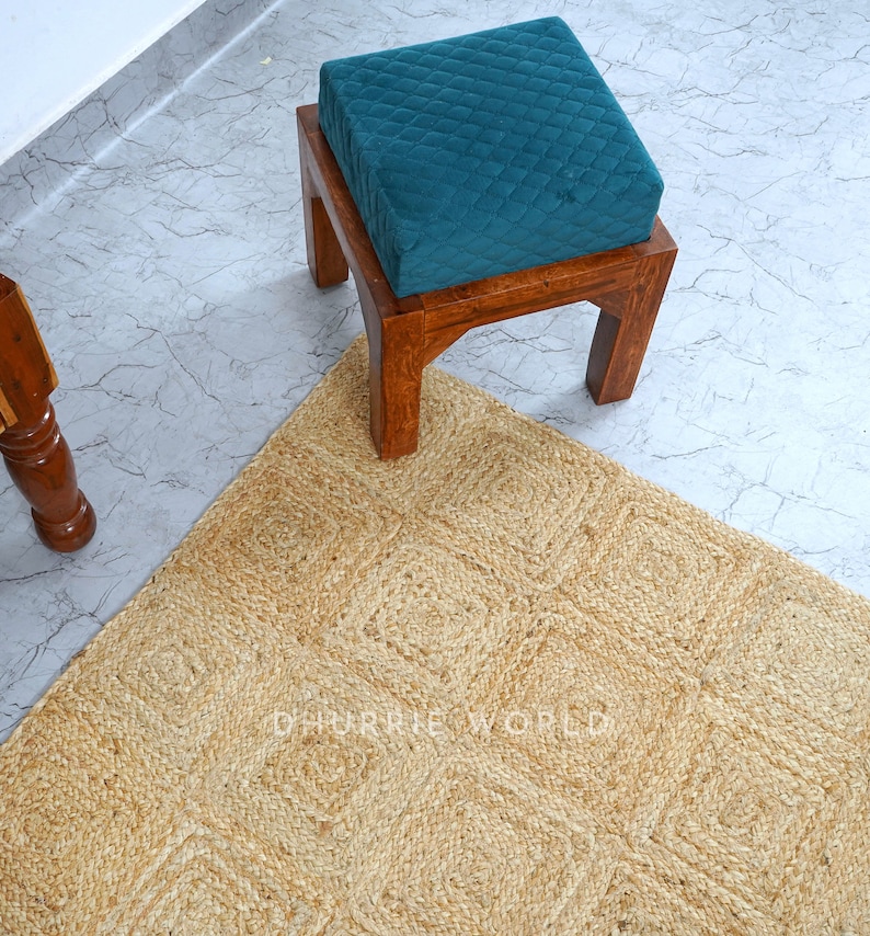 Puede incluir: Un coj&iacute;n de terciopelo turquesa se sienta sobre un taburete de madera con una alfombra de yute natural debajo. La alfombra tiene un patr&oacute;n tejido de cuadrados.