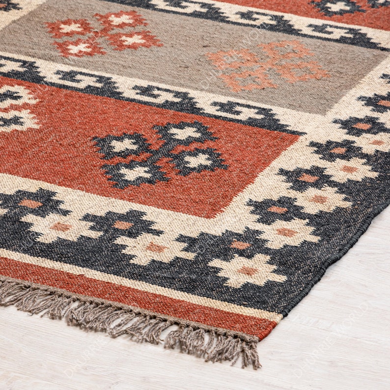 May include: A hand-woven rug with a geometric pattern in shades of red, black, white, and beige. The rug has a fringe border.