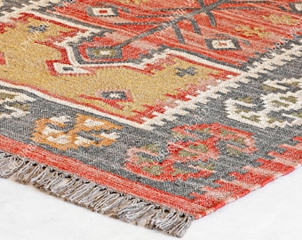 May include: A red, grey and beige patterned rug with fringe on the edges. The rug has a geometric design with a tribal feel.