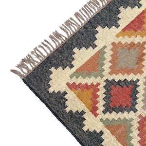 May include: A close-up of a colorful rug with a geometric pattern. The rug is made of natural fibers and has a fringe border. The colors in the rug are black, white, red, orange, yellow, and green.