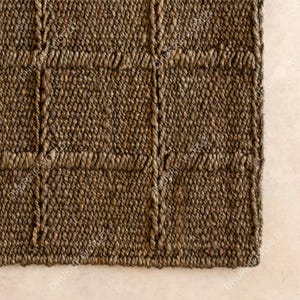 Puede incluir: Primer plano de una alfombra marr&oacute;n tejida con un patr&oacute;n de cuadr&iacute;cula. La alfombra est&aacute; hecha de fibras naturales, con una superficie texturizada y una paleta de colores neutros. El dise&ntilde;o presenta una serie de cuadrados y rect&aacute;ngulos.