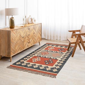 May include: A rectangular rug with a geometric pattern in shades of red, black, beige, and gray. The rug has a fringe border and is laid out on a wooden floor in a room with a wooden dresser and a chair.