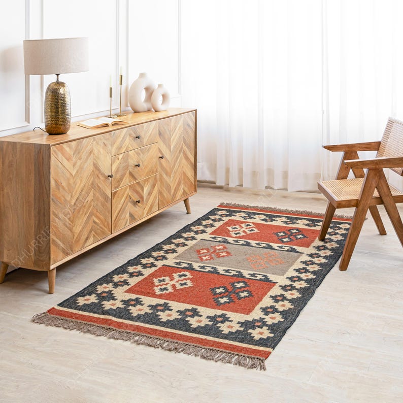 May include: A rectangular rug with a geometric pattern in shades of red, black, beige, and gray. The rug has a fringe border and is laid out on a wooden floor in a room with a wooden dresser and a chair.