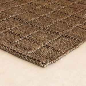 Puede incluir: Primer plano de una alfombra tejida marr&oacute;n con un patr&oacute;n geom&eacute;trico en forma de cuadr&iacute;cula. La alfombra est&aacute; hecha de una fibra natural, posiblemente yute o sisal, y tiene un aspecto texturizado. El color neutro y el dise&ntilde;o sencillo la hacen adecuada para varios estilos de interior.