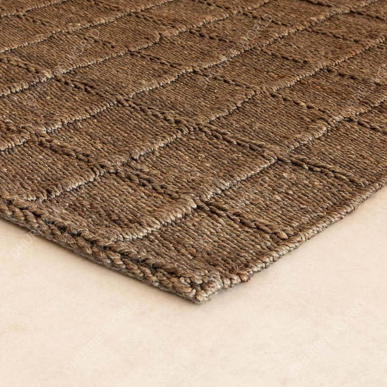 Puede incluir: Primer plano de una alfombra tejida marr&oacute;n con un patr&oacute;n geom&eacute;trico en forma de cuadr&iacute;cula. La alfombra est&aacute; hecha de una fibra natural, posiblemente yute o sisal, y tiene un aspecto texturizado. El color neutro y el dise&ntilde;o sencillo la hacen adecuada para varios estilos de interior.