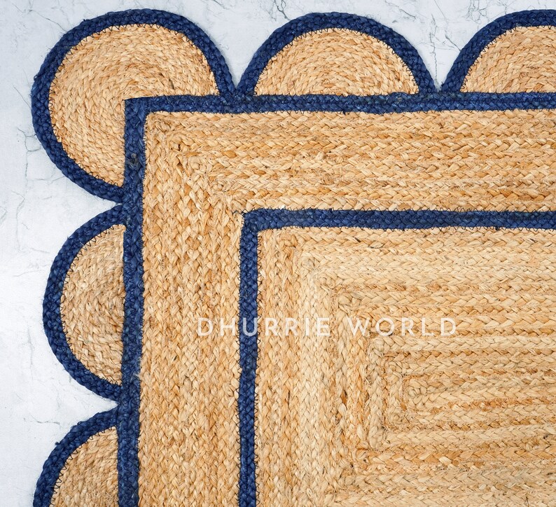 May include: A natural fiber rug with a square shape and a scalloped border. The rug has a dark blue trim around the edges and a smaller square in the center. The text "DHURRIE WORLD" is visible in the center of the rug.