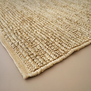 May include: A rectangular area rug featuring a thick, hand-woven texture made from natural tan fibers. The rug displays a consistent, repeating looped weave pattern across its surface, finished with a clean, straight edge along the border.