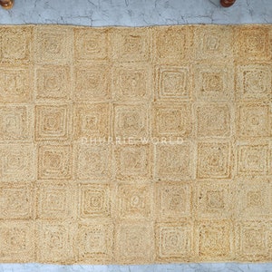 Puede incluir: Una alfombra de fibra natural con un patr&oacute;n cuadrado. La alfombra est&aacute; hecha de yute tejido y tiene un color marr&oacute;n claro.