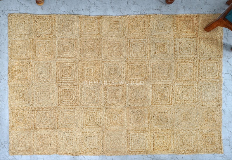 Puede incluir: Una alfombra de fibra natural con un patr&oacute;n cuadrado. La alfombra est&aacute; hecha de yute tejido y tiene un color marr&oacute;n claro.