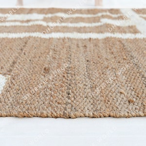 May include: Close-up of a natural fiber rug with a white geometric pattern. The rug is made of jute and has a natural, earthy tone.