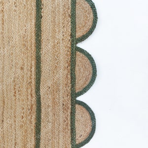 K&ouml;nnte beinhalten: Ein Naturfaser-Teppich mit gr&uuml;nem Rand und einem gewellten Saum. Der Teppich ist aus Jute und hat eine nat&uuml;rliche, erdige Farbe.