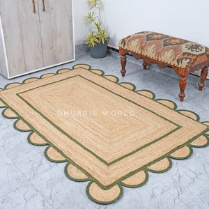 May include: A natural fiber rug with a green border and scalloped edges. The rug is made of jute and has a natural, earthy color. The rug is placed on a tiled floor and a small wooden bench with a patterned cushion is in the background. "DHURRIE WORLD" is printed on the rug.