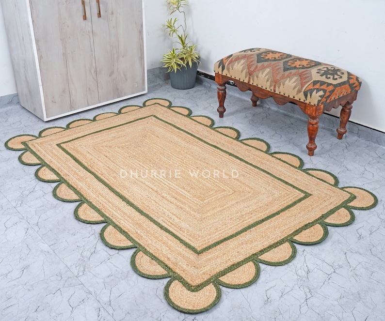 May include: A natural fiber rug with a green border and scalloped edges. The rug is made of jute and has a natural, earthy color. The rug is placed on a tiled floor and a small wooden bench with a patterned cushion is in the background. "DHURRIE WORLD" is printed on the rug.