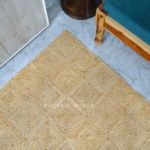 Puede incluir: Una alfombra de fibra natural con un patr&oacute;n cuadrado en color beige claro. La alfombra est&aacute; hecha de yute y tiene una superficie texturizada.