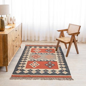 May include: A red, black, and beige patterned rug with a geometric design. The rug has a fringe border and is laid out on a wooden floor.
