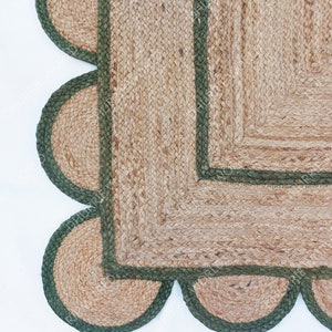 K&ouml;nnte beinhalten: Ein quadratischer Jute-Teppich mit gr&uuml;nem Rand und einem gewellten Saum. Der Teppich ist aus Naturfasern gefertigt und hat einen rustikalen Look.