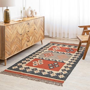 May include: A hand-woven rug with a geometric pattern in shades of red, black, and beige. The rug has a fringe border and is made of natural fibers.