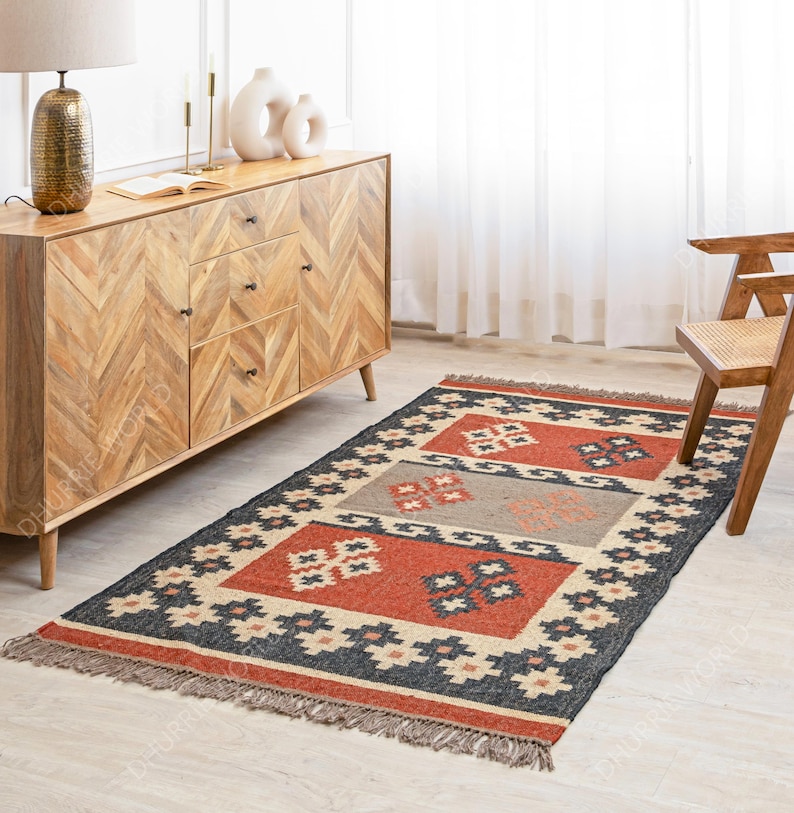 May include: A hand-woven rug with a geometric pattern in shades of red, black, and beige. The rug has a fringe border and is made of natural fibers.