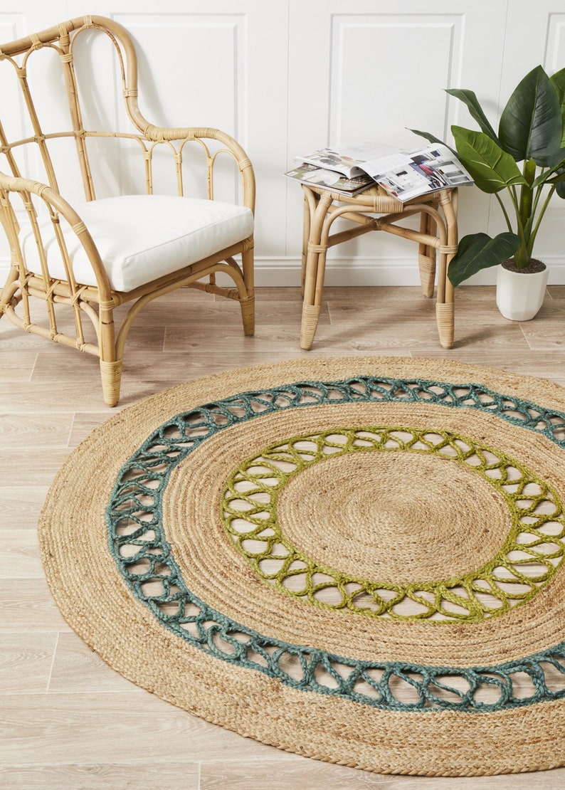 May include: Round jute rug with a natural tan color, featuring a teal and green decorative border. The rug is placed on a light wood floor, with a rattan chair and side table in the background.