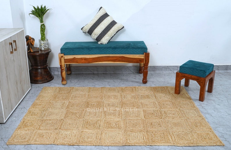 Puede incluir: Una alfombra de yute natural con un patr&oacute;n cuadrado de color marr&oacute;n claro. La alfombra se coloca delante de un banco de madera con un coj&iacute;n turquesa y un peque&ntilde;o taburete de madera con un coj&iacute;n turquesa.