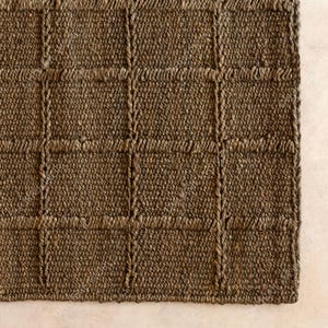 Puede incluir: Primer plano de una alfombra marr&oacute;n tejida con un patr&oacute;n de cuadr&iacute;cula. La alfombra est&aacute; hecha de fibras naturales, con una superficie texturizada y una paleta de colores neutros. El dise&ntilde;o presenta una serie de cuadrados y rect&aacute;ngulos.