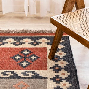May include: A hand-woven rug with a geometric pattern in shades of red, black, white, and beige. The rug has a fringe border and is partially covered by a wooden chair with a woven cane seat.