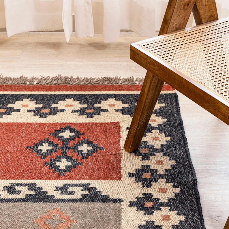 May include: A hand-woven rug with a geometric pattern in shades of red, black, white, and beige. The rug has a fringe border and is partially covered by a wooden chair with a woven cane seat.