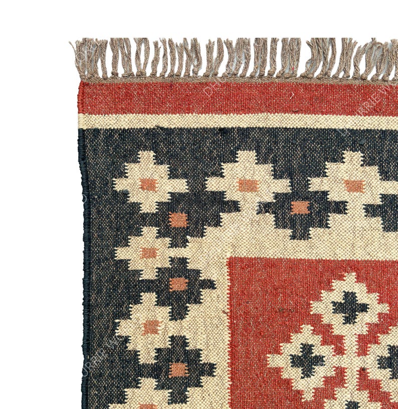May include: A handwoven rug with a geometric pattern in shades of red, black, and cream. The rug has a fringe border.