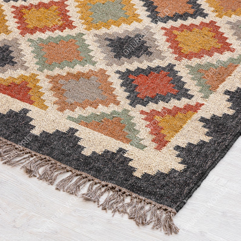 May include: A close-up of a woven rug with a geometric pattern in shades of brown, orange, green, and black. The rug has a fringe border.