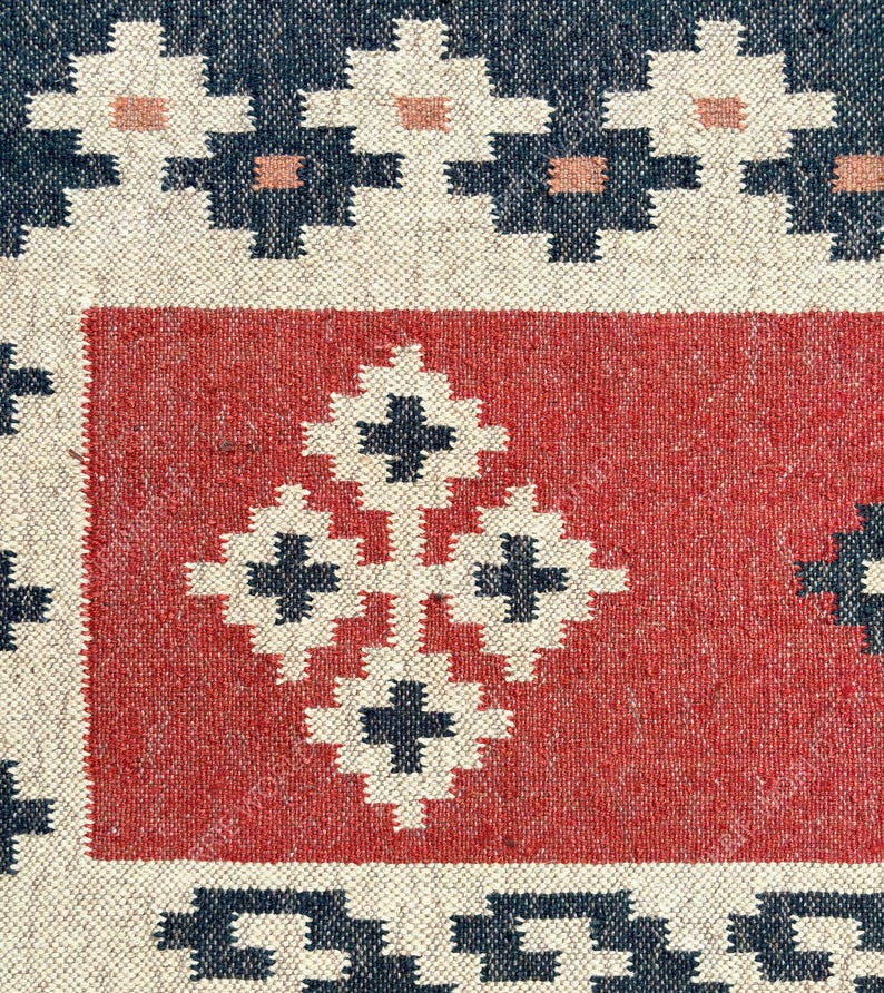May include: A close-up of a red and white woven rug with a geometric pattern. The rug has a dark blue border and a central design of four white squares with black crosses.