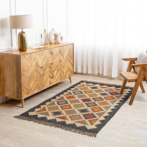 May include: A black and brown patterned rug with fringe edges. The rug is laid out on a wooden floor in a living room setting with a wooden dresser and a chair.