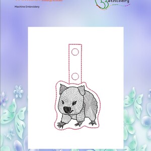 May include: A machine embroidery design of a gray wombat with a red loop for a key fob. The design is on a white background with a floral border.