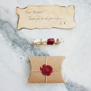 May include: A gift set with a vintage aesthetic. The set includes a corrugated cardboard box tied with twine and sealed with a red wax stamp. A rolled parchment scroll is also sealed with red wax. A handwritten note reads "Dear Margaret. Thank you for all your help. H.N."