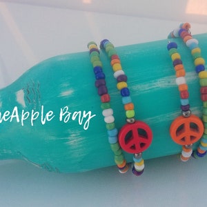 May include: Two beaded bracelets with a peace sign charm. The bracelets feature colorful beads in a rainbow pattern and a red or orange peace sign charm. The bracelets are displayed on a turquoise painted surface. The text "PineApple Bay" is visible.