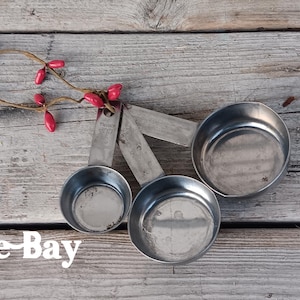 May include: Three stainless steel measuring cups of varying sizes are arranged on a weathered wooden surface. A sprig of red berries adds a touch of color. The text "PineApple Bay" is displayed in white.